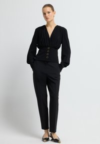 Blusa nera con scollatura a V profonda, spalle plissettate e vita aderente accentuata da bottoni dorati, abbinata a pantaloni neri su misura e scarpe nere.