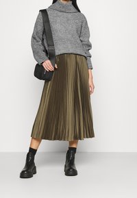 Grauer Strickpullover mit hohem Kragen, kombiniert mit einem plissierten, olivgrünen Midirock. Schwarze Stiefeletten und eine schwarze Schultertasche vervollständigen den Look.