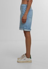 Jupe en denim bleu clair, longueur genoux avec un ourlet roulé. Associée à des baskets blanches avec des accents verts et des détails écrits à la main.