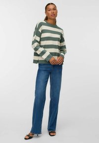 Donna in piedi che indossa un maglione a righe verdi e bianche, jeans blu e sandali neri con la punta aperta su uno sfondo bianco uniforme.