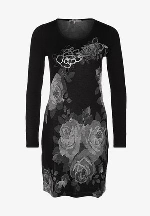 Mujer vistiendo un vestido negro de manga larga con un patrón floral plateado, medias negras, zapatos de punta negros y sosteniendo un bolso de mano negro.