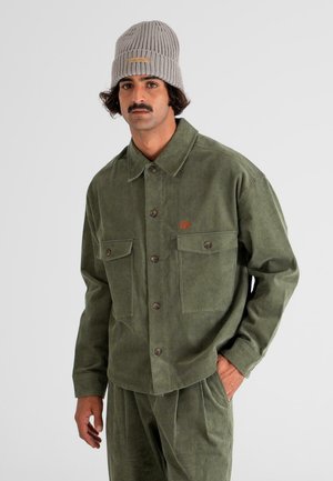 Man wearing olive green corduroy jacket and pants with front pockets and a light gray knit beanie against a plain background.