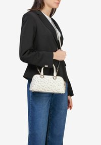 Hvid håndtaske med prikket tekstur, rund form, dobbelte håndtag og en aftagelig rem. Båret med en sort blazer og blå jeans.