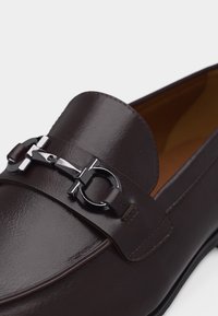 Mocassin en cuir marron foncé avec détail mors en argent et finition lisse, montrant le dessus avant et une partie de la semelle.