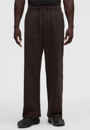 Uomo con pantaloni della tuta larghi marrone scuro con vita elastica e coulisse, abbinati a scarpe sportive nere.