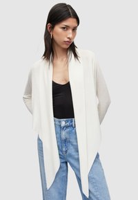 Langer, offener Cardigan in hellem Creme, aus weichem Stoff; verfügt über ein drapiertes Design und eine gerippte Textur. Kombiniert mit einem dunklen Oberteil und Jeans.