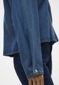 Modra jeans srajca z dolgimi rokavi, ki ima zapenjanje na manšetah in neobdelan rob. Tkanina ima subtilno teksturo, dopolnjeno z kontrastnim šivanjem.