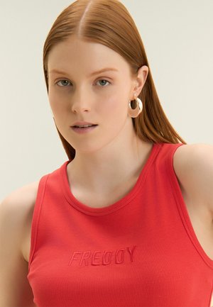 Femme aux longs cheveux roux et aux yeux verts portant un haut rouge sans manches avec "FREDDY" brodé et de grandes boucles d'oreilles en or.