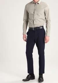 Man med beige skjorta med knappar, marinblå kostymbyxor med svart bälte, svarta strumpor och svarta klädskor, står mot en enfärgad bakgrund.