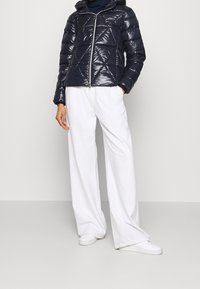 Marinblå vadderad pufferjacka med huva, blank yta, dragkedjestängning och diamantmönster. Kombinerad med vita vida byxor och vita sneakers.