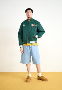 Grøn satin bomberjakke med hvid- og grønstribede ribbede manchetter, broderede grafikker, lagdelt over en gul skjorte og denimshorts.