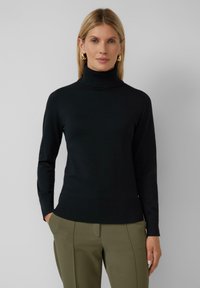 Zwarte coltrui in geribbeld textiel met lange mouwen en een aansluitend ontwerp. Gepaard met olijfgroene op maat gemaakte pantalons.