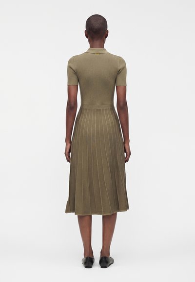Femme portant une robe en maille côtelée vert olive avec manches courtes et jupe plissée, debout de dos, sur fond blanc.