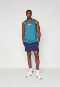 Turkis ermeløs singlet med oransje logo; kombinert med marineblå shorts og hvite treningssko; avslappet sporty stil.
