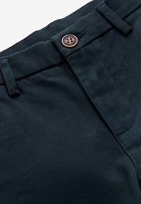 Pantaloni bleumarin închis cu un nasture metalic vizibil gravat cu inscripția „Authentic Next Dude” și o buclă de curea deasupra taliei.