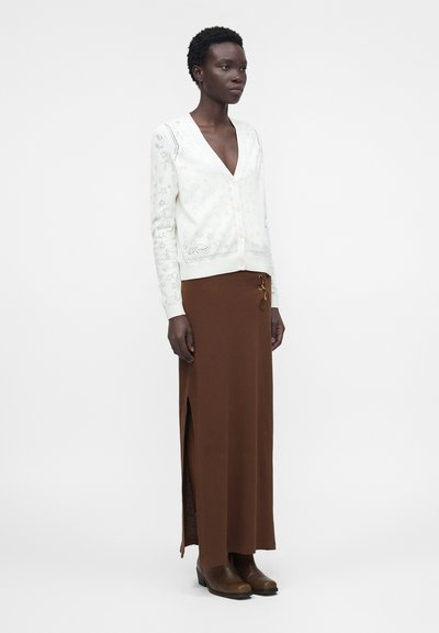 Γυναίκα που φορά λευκή ζακέτα με κουμπιά και floral σχέδια, καφέ μακριά φούστα με πλευρικό άνοιγμα και καφέ μποτάκια μέχρι τον αστράγαλο, στέκεται μπροστά σε λευκό φόντο.