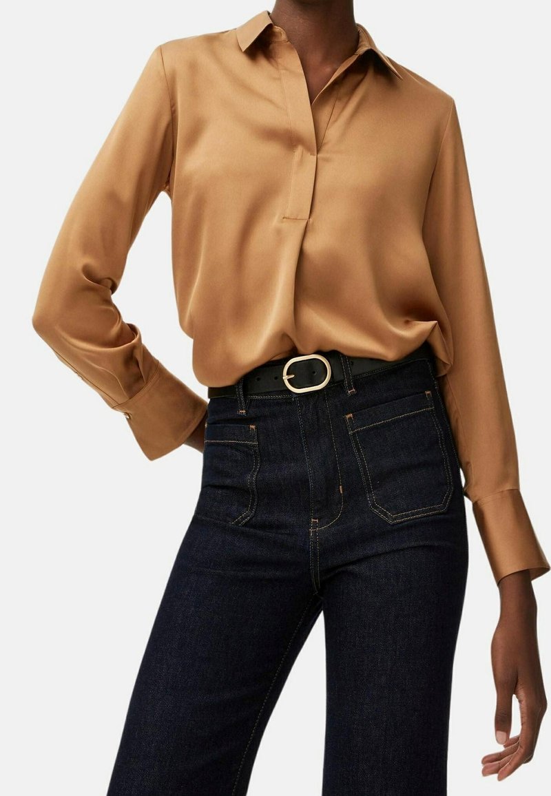 Blusa beige in satin con pattina a bottoni, maniche lunghe e dettagli ai polsini, abbinata a jeans in denim scuro a vita alta e una cintura nera.