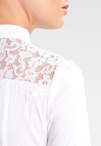 Bílá košile s krajkovým zadním panelem, která má květinový vzor a texturované detaily. Límec je klasický s knoflíkovým zapínáním vpředu.