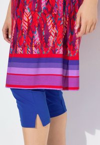 Červená tunika s vzorem pírků v několika barvách, doplněná modrými šortkami s bočními rozparky pro snadný pohyb.