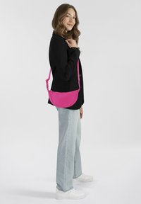 Borsa a tracolla rosa con forma arrotondata, materiale in tessuto e tracolla regolabile. Indossata da una persona con una giacca nera e jeans chiari.