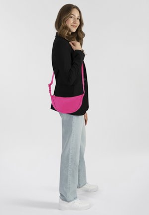 Borsa a tracolla rosa con forma arrotondata, materiale in tessuto e tracolla regolabile. Indossata da una persona con una giacca nera e jeans chiari.