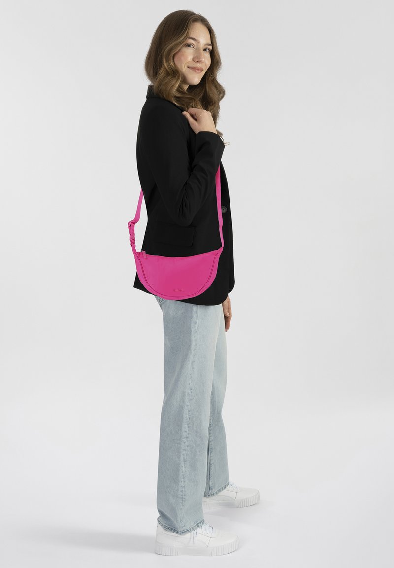 Borsa a tracolla rosa con forma arrotondata, materiale in tessuto e tracolla regolabile. Indossata da una persona con una giacca nera e jeans chiari.