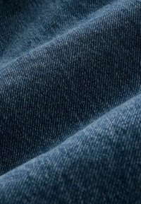 Denim tkanina srednje plave boje, s tkanjem u tvid stilu i vidljivim detaljima šivanja, prikazuje teksturu i dubinu.
