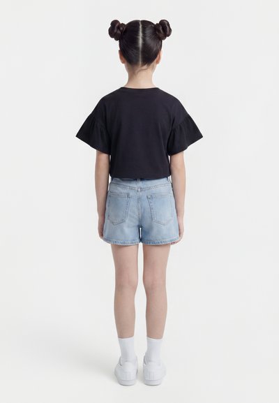 Κορίτσι με δύο κότσους, που φοράει μαύρο τοπ με βολάν στα μανίκια, ανοιχτόχρωμο τζιν σορτς, λευκές κάλτσες και λευκά αθλητικά παπούτσια, στέκεται πλάτη.