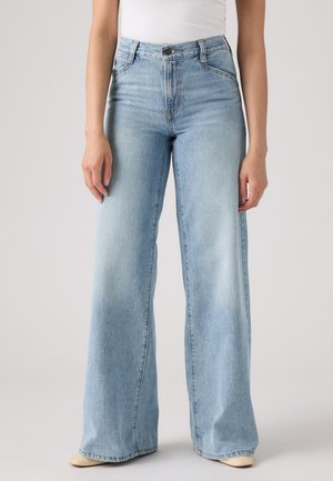 Mujer que lleva vaqueros de tiro alto de pierna ancha de color azul claro con botón y bolsillos delanteros, combinados con zapatos beige y una camiseta blanca.