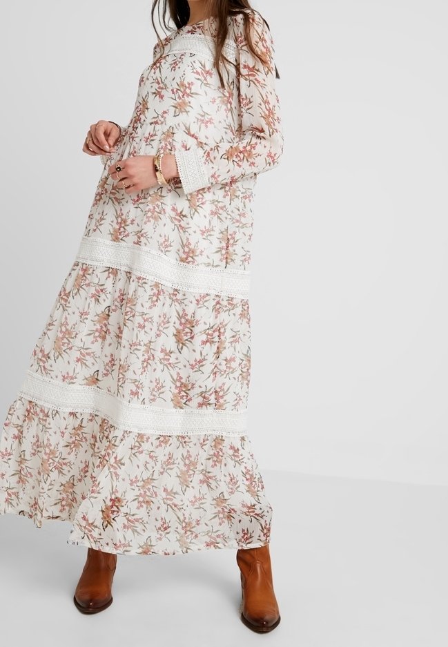 Une longue robe florale blanche avec des motifs roses et verts, des accents en dentelle au niveau des manches et de la taille, portée avec des bottines brunes à la cheville.