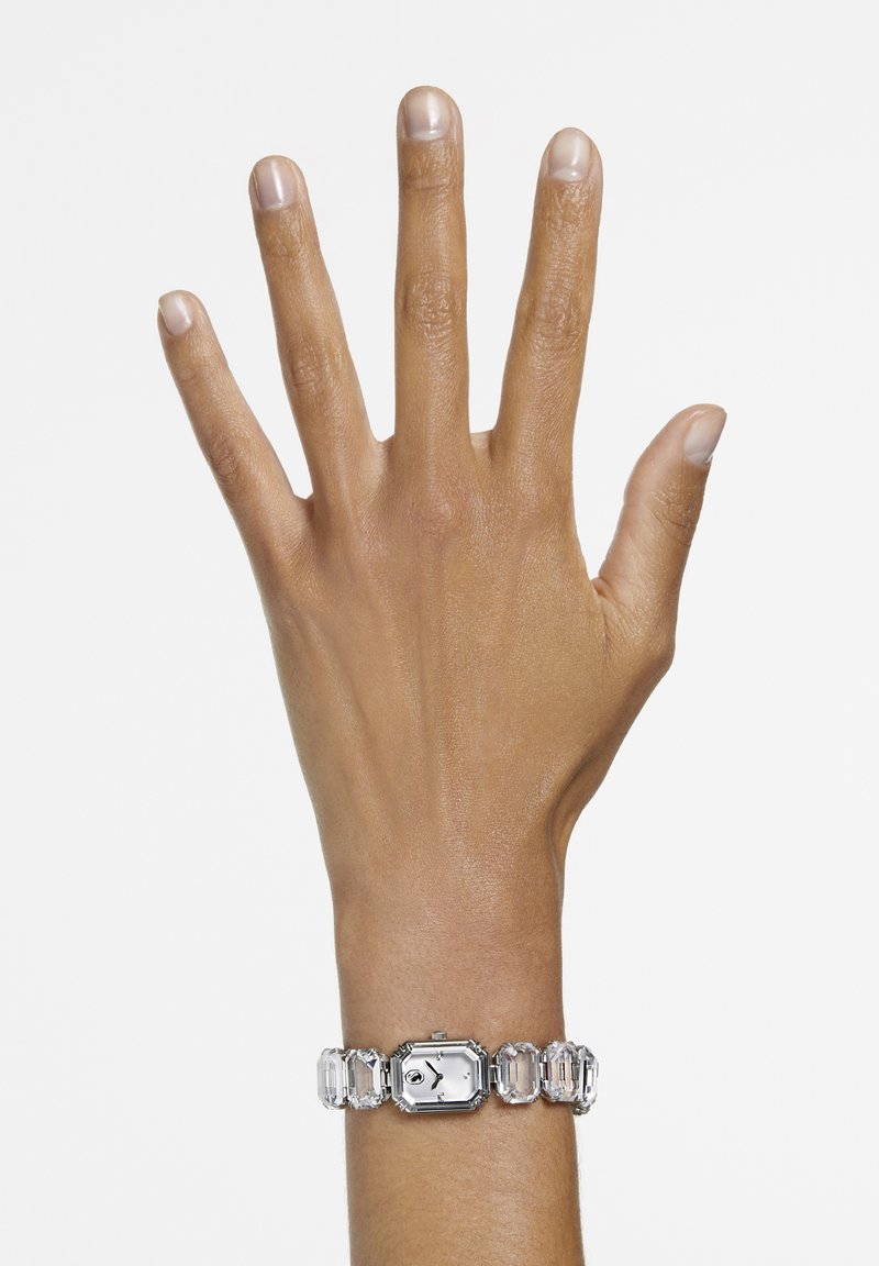 Reloj plateado con esfera octagonal y acentos de cristal rectangular, llevado en una mano, mostrando un diseño elegante y detalles claros.
