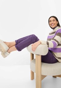 Maglione lavorato a maglia con strisce viola, marroni e bianche, abbinato a pantaloni viola e scarpe platform beige. Seduto su una sedia rotonda imbottita.