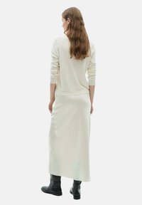Biały sweter z dzianiny w połączeniu z długą, zwiewną białą spódnicą. Modelka nosi czarne botki za kostkę. Gładka faktura, minimalistyczny design.