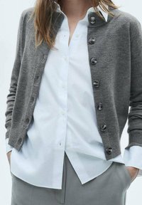 Graue Strickjacke aus weichem Material mit Knopfleiste und dunklen Knöpfen, getragen über einem weißen Hemd mit Kragen. Schlichtes Design.