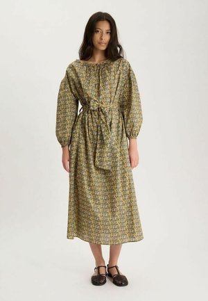 Femme portant une robe jaune à manches longues et longue jusqu’aux chevilles avec un motif floral et une ceinture nouée à la taille, associée à des chaussures noires tissées.