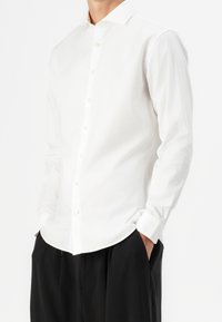 Camicia bianca con colletto stretto, maniche lunghe e orlo curvo. Realizzata in tessuto di cotone liscio, presenta un design a vestibilità classica.