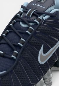 Tuvumā redzams tumši zils Nike Shox apavu modelis ar gaismas zila akcentiem, pelēku zīmolā Swoosh un redzamu atsperu pēdas amortizāciju.
