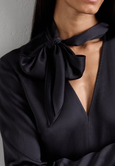 Blusa nera realizzata in tessuto liscio, con scollatura a V e un grande fiocco decorativo al collo. Il design presenta linee pulite e uno stile minimalista.
