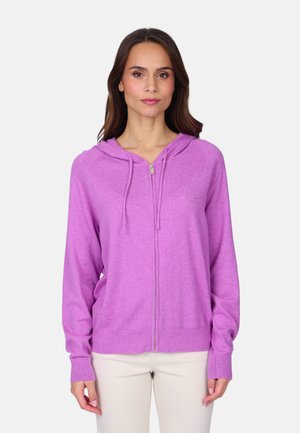 Femme portant un sweat à capuche zippé violet clair avec des cordons, debout devant un fond clair uni, les mains détendues le long du corps.
