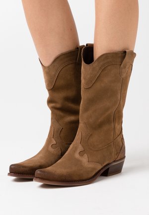 Botas de vaquero de ante marrón con punta afilada, parte superior en forma de ondas y patrones de costura decorativos. Cuentan con un tacón bajo y apilado.