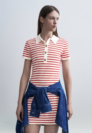 Kvinde iført en rød og hvidstribet kortærmet kjole med krave og knapper, blå denimjakke bundet om livet, kigger til siden.