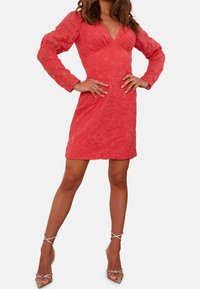 Korallklänning med långa, strukturerade ärmar, en V-ringning och blommönster. Kombineras med klara, remmade klackar som framhäver en kroppsnära silhuett.