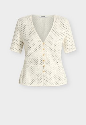 Kvinde iført en creme kortærmet striktop, brune vidde bukser, sorte sandaler og holder en beige håndtaske med den ene hånd i lommen.