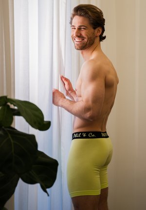 Pánské boxerky ve výrazné žluté barvě z bavlny s černým logem na pase. Model stojí u okna lemovaného jemnými závěsy.