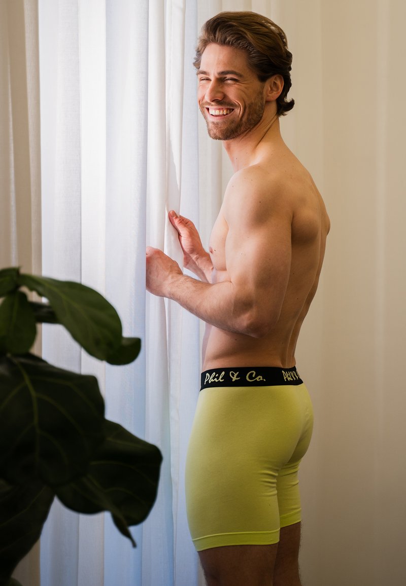 Boxer briefs para hombres en algodón amarillo brillante con una cintura con logo negro. El modelo está de pie junto a una ventana delimitada por cortinas transparentes.