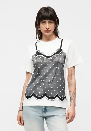 Persoană cu păr închis la culoare, purtând un tricou alb peste care are un top negru transparent cu buline și blugi deschis la culoare, stând pe un fundal simplu.