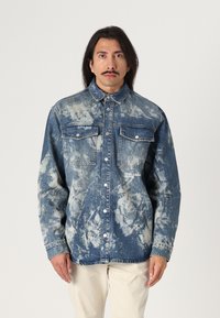Denim-paita, jossa on sinivalkoinen tie-dye-kuvio, jossa on kaksi rintataskua, napit ja logo vasemmalla puolella.