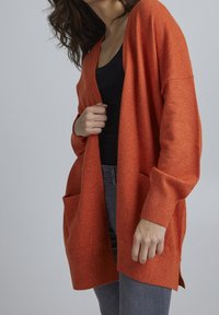 Přehnaně velký oranžový cardigan z měkkého materiálu s hlubokým výstřihem do V, žebrovanými manžetami a dvěma předními kapsami. Nenošený přes černý top a šedé džíny.