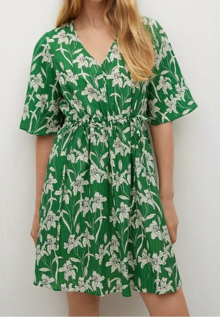 Robe courte verte avec motif floral blanc, col en V, manches bouffantes et taille cintrée, portée par une personne aux longs cheveux blonds.