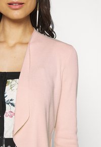 Cardigan rosa chiaro con una morbida texture a maglia, caratterizzato da un design aperto, indossato su una maglietta a fantasia floreale.